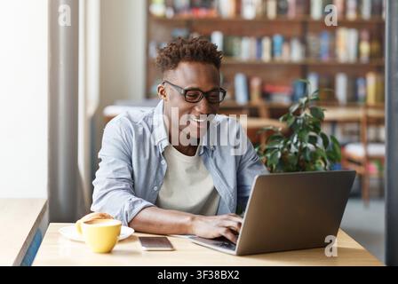 Zufriedener schwarzer Mann arbeitet an einem Laptop im Café, konzentriert und produktiv, Geschäftskonzept Stockfoto