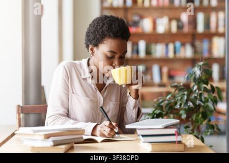 Konzentrierte schwarze Frau schreibt in ein Notizbuch, während sie in einem gemütlichen Café Kaffee trinkt Stockfoto