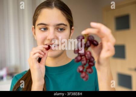 Junge Frau isst rote Trauben im Innenraum. Junge Frau in einem blaugrünen Hemd hält einen Bunch roter Trauben und führt eine Traube zum Mund in sanftem Licht Stockfoto