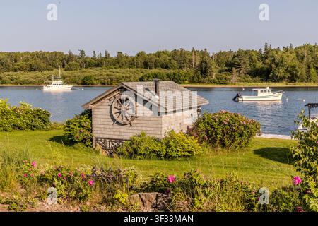 Ein kleiner Gartenschuppen, geschmückt mit einem Schiffssteuerrad, steht am Ufer der Water Street in Baddeck, Nova Scotia, Kanada, mit zwei Fischerbooten auf dem Bras d’Or Lake im Hintergrund Stockfoto