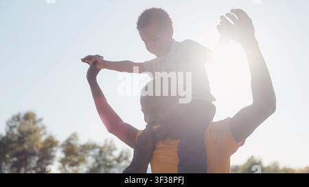 Ein alleinerziehender Vater gibt seinem Sohn eine Fahrt auf seinen Schultern, während sie bei Sonnenuntergang gemeinsam die Natur genießen. Stockfoto