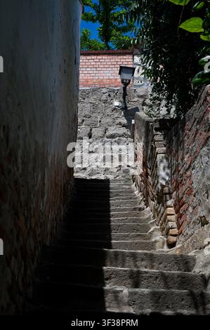 Schmale historische Steintreppe, die zwischen alten Ziegel- und Steinmauern hinaufführt, mit einer antiken Straßenlaterne in Szentendre, Ungarn. Stockfoto