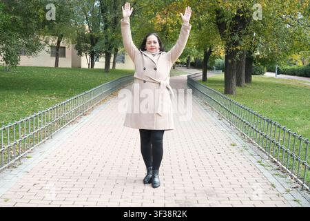 Frau steht auf gepflastertem Weg mit erhobenen Armen. Sie trägt einen beigen Mantel und schwarze Stiefel. Bäume säumen den Gehweg hinter ihr. Grünes Gras säumt den Weg. Stockfoto