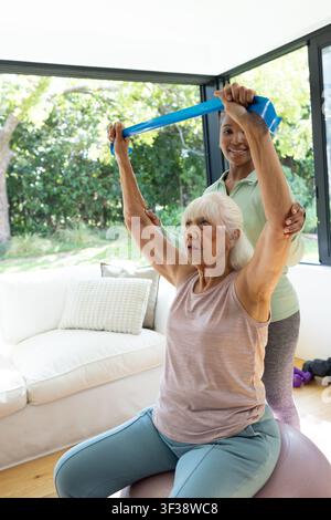 Vielfältige weibliche Betreuerin führt eine ältere Frau, die auf einem Stabilitätsball sitzt und ein blaues Band verwendet, zu Hause Stockfoto