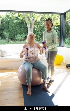 Seniorin sitzt zu Hause auf einem Ball und benutzt lila Gewichte als weibliche Trainerin in Sportkleidung, die sie beobachtet Stockfoto