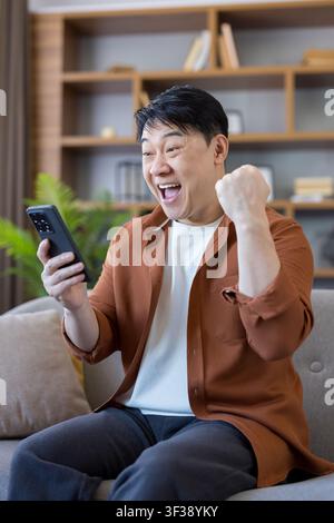 Fröhlicher Asiate sitzt auf einem Sofa, hebt triumphierend die Faust und schaut auf sein Smartphone, feiert einen Sieg oder gute Nachrichten, die über das Gerät empfangen wurden Stockfoto