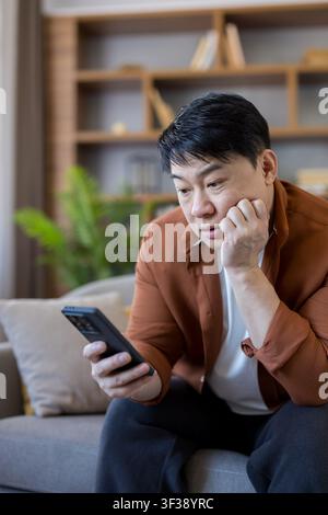 Reifer Mann sitzt zu Hause auf dem Sofa, betrachtet nachdenklich sein Smartphone, verbindet sich mit digitalem Inhalt, überlegt Online-Informationen, fühlt sich überrascht, besorgt oder denkt über eine Nachricht nach Stockfoto