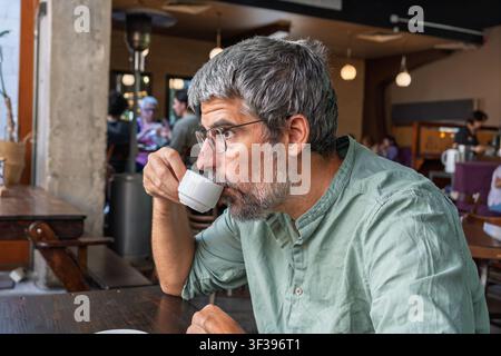 Reifer Mann trinkt Kaffee aus einer kleinen weißen Tasse und entspannt sich in einem gemütlichen Café mit verschwommenen Menschen im Hintergrund Stockfoto
