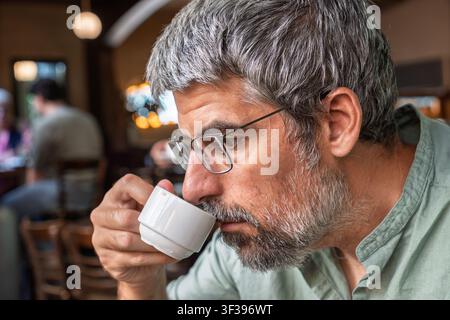 Mann mit grauen Haaren und Bart, der Espresso aus einer weißen Tasse trinkt und sich in einer authentischen Café-Atmosphäre entspannt Stockfoto