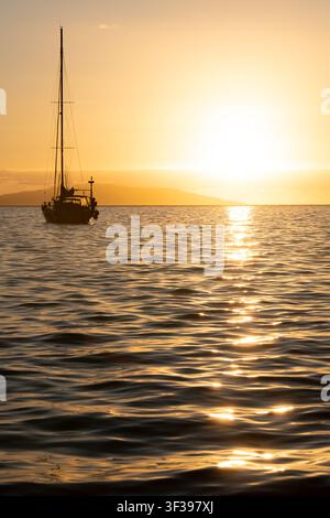 Ein Segelboot im Silhouettenschnitt vor Anker auf einem ruhigen goldenen Meer, während die Sonne hinter einer fernen Insel vor La Gomera, Kanarische Inseln, Spanien, untergeht Stockfoto