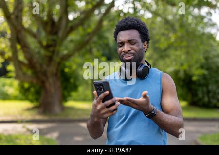 Erwachsener Mann in Sportkleidung trägt Kopfhörer, blinzelt und zeigt Verärgerung, während er in einer Parkumgebung im Freien mit seinem Mobiltelefon interagiert und auf technische Probleme oder eine verwirrende Benutzeroberfläche stößt Stockfoto