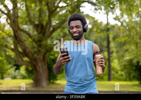 Zufriedener junger Mann mit Kopfhörern, der ein Smartphone und eine Wasserflasche hält, durch Inhalte scrollt und lächelt, während er nach dem Sport in einem grünen Park steht Stockfoto