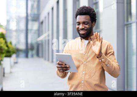 Junger afrikanischer Mann im Freien, der lächelt und winkt, während er ein digitales Tablet hält, und an einem fröhlichen Videoanruf für Remote-Arbeit, Networking oder virtuelle Besprechungen in der Stadt teilnimmt Stockfoto