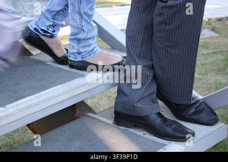 Füße auf den grauen Stufen. Füße in Schuhen. Perilla. Schwarze Schuhe. Schwarze Ballettschuhe. Graue Hosen. Blaue Jeans. Frauen- und Männerbeine Stockfoto