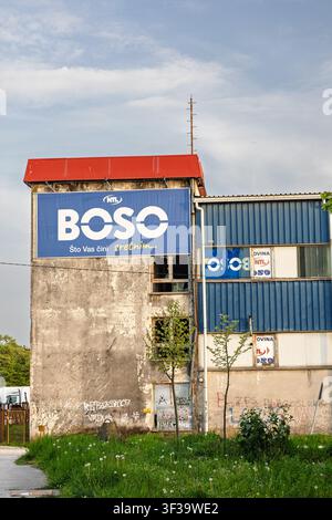 Vinkovci, Kroatien - 15. April 2026: Langes, niedriges Industriegebäude, blau gestrichen mit Graffiti entlang des Betonfundaments, das hinter einem grasbewachsenen Gelände unter einem Stockfoto