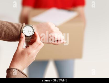 Lieferung pünktlich. Weibliche Hände mit Uhr prüfen die Zeit, Nahaufnahme, Kurier mit Kiste und Tablet Stockfoto