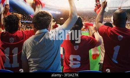 Nahaufnahme von glücklichen Fußballfans in roten Trikots, die mit Vuvuzelas singen und in den Stadiontribünen laufen. Lächelnde Fußballfans blasen Lufthörner, um ihr Team während des Spiels anzufeuern Stockfoto