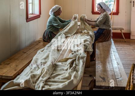 Zwei weibliche Demonstrantinnen in traditioneller Kleidung falten ein handgewebtes Wolltuch auf einer Tischbank im Highland Village Museum | Baile nan Gàidheal in Iona, Nova Scotia, Kanada. In einem als Waulking (Gälisch: luadh) bekannten Prozess wurde frisch gewebter Tweed durch rhythmisches Reiben und Falten von Hand verdichtet und verfilzt, traditionell begleitet von Arbeitssongs Stockfoto