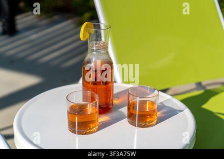 Nahaufnahme von Eisteegläsern und Karaffe auf weißem Terrassentisch neben einem grünen Loungesessel Stockfoto