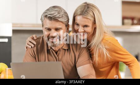 Zufriedenes reifes kaukasisches Paar lächelt zusammen am Laptop in moderner Küche, Technologie-Konzept Stockfoto