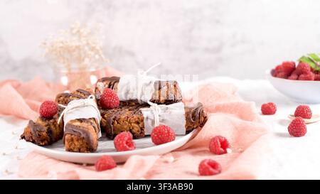 Hausgemachte Granola-Riegel, die mit dunkler Schokolade beträufelt, in Pergamentpapier eingewickelt und mit frischen Himbeeren garniert sind, werden auf einem weißen Teller serviert über Stockfoto
