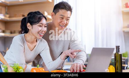 Lächelndes asiatisches Paar kocht gemeinsam in einer modernen Küche und nutzt einen Laptop für Rezepte Stockfoto