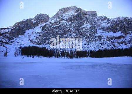 Schneebedeckte Berge mit gefrorenem See im Vordergrund und Wald dahinter in der Dämmerung, Pragser Wildsee, Lago di Prags, gefrorener Bergsee im Winter Stockfoto