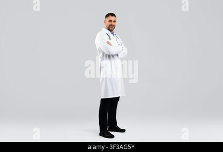 Fröhlicher Arzt mittleren Alters, der mit gefalteten Armen posiert und auf die Kamera blickt, auf einem hellen Hintergrund steht, volle Körperlänge Stockfoto