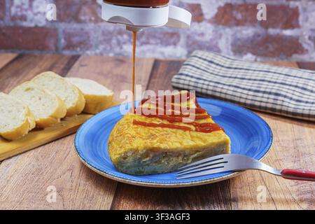 Scheibe traditionelles spanisches Omelett mit Ketchup auf einem blauen Teller, serviert mit frischem Brot auf einem rustikalen Holztisch Stockfoto