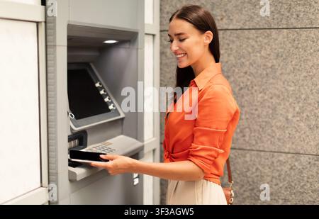 Lächelnde Dame, die das Telefon zum Geldautomaten bringt und Bargeld draußen abzieht Stockfoto