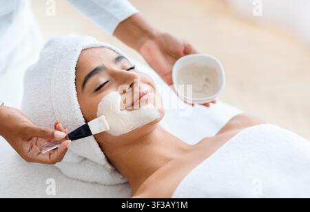 Kosmetologe, der Tonmaske auf das Gesicht einer indischen Frau aufträgt Stockfoto