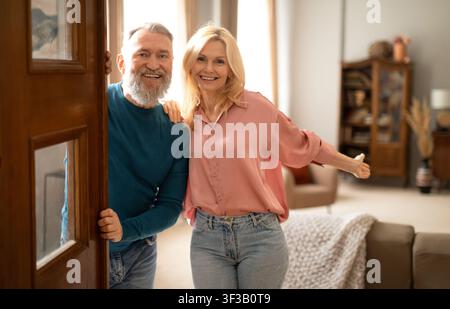Glückliches Seniorenpaar, das eine offene Tür hält und drinnen auf die Kamera schaut Stockfoto