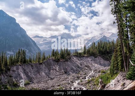 Majestätische Berge erheben sich hinter einem rauen, felsigen Tal. Wolken ziehen über schneebedeckten Gipfeln und dichten immergrünen Bäumen. Ein sich windender Bach fließt durch gefallene S Stockfoto