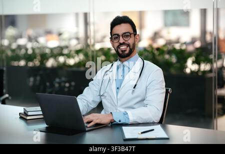 Lächelnd attraktiver junger arabischer Arzt mit Bart in Brille und weißem Mantel, am Arbeitsplatz mit Laptop arbeiten Stockfoto