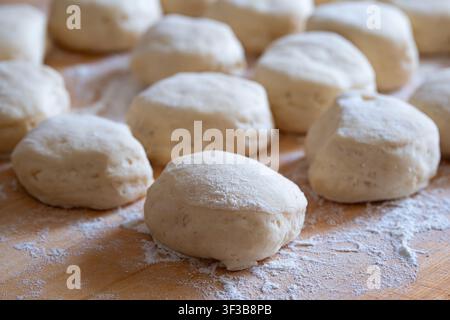 Eine Nahaufnahme, die einen einzelnen rohen runden Teigling im scharfen Fokus zeigt, umgeben von anderen mit Mehl bestäubten Teigstücken, die auf einer Holzschneideplatte ruhen Stockfoto