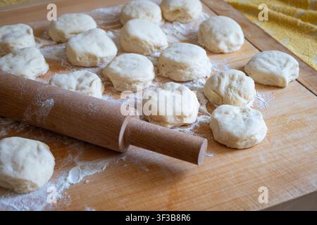 Eine seitliche Nahaufnahme, die eine klassische hölzerne Nudelrolle zeigt, die zwischen rohen runden Hefteigstücken liegt, die mit Mehl bestäubt sind, auf einem großen Holzschneidebrett Stockfoto