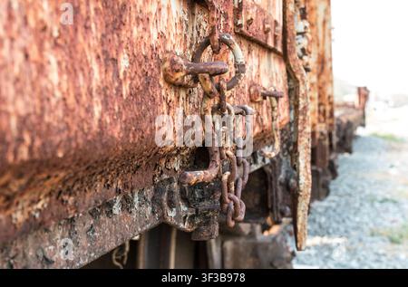Nahaufnahme einer rostigen Kette und eines Ringverschlusses an einem alten Güterwaggon mit einer Reihe von Waggons im Hintergrund Stockfoto