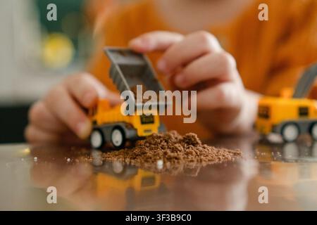 Dynamisches Kleinkind bewegt zwei gelbe Spielzeugkipplaster, wodurch Sand und Schmutz auf dem Tisch verschüttet werden. Verspieltes Kind schiebt Fahrzeuge in aktivem, energischem Spiel und zeigt den Prozess. Der Kleine erforscht die Bewegung beim Entladen von Erde Stockfoto