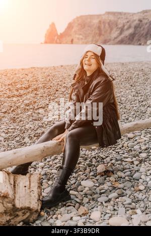 Frau Strand Sonnenuntergang glückliche junge Dame, die auf einem Treibholzstamm an einem Kiesufer während eines warmen goldenen Sonnenuntergangs sitzt Stockfoto