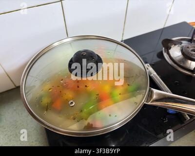 Frische gemischte Gemüse dampfen unter Glasdeckel in Edelstahlbratpfanne Stockfoto