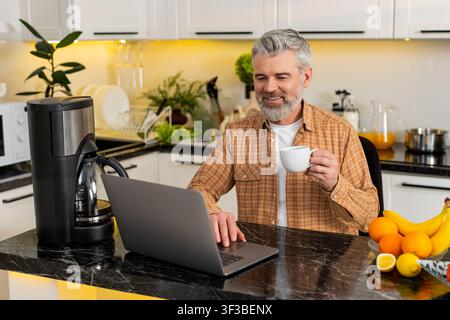 Ein Mann mittleren Alters arbeitet an einem Laptop in der Küche und trinkt heißen Kaffee, während er E-Mails für freiberufliche Fernarbeit liest. Reifer Kerl fühlt sich glücklich produktiv während der Pause und hält weiße Tasse in der modernen Küche. Stockfoto