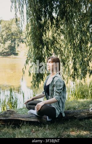 Frau meditiert am See unter Weidebaum, Waldbaden, Atemübung, Erdungstechniken, mentale Klarheit Stockfoto