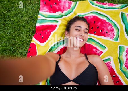 Fröhliche junge Frau lächelt, liegt auf einem bunten Wassermelonenhandtuch im Park und fängt einen Moment sommerlicher Entspannung im Urlaub ein Stockfoto