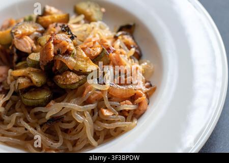 Extremer Makro-Nahaufnahme von Shirataki-Nudeln mit geflocktem Lachs, sautiertem Zucchini und karamellisierten Zwiebeln auf einem weißen Teller Stockfoto