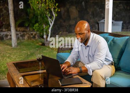 Lächelnder schwarzer Geschäftsmann, der nachts auf einer Außenterrasse remote mit einem Laptop arbeitet, neben einem Glas Rotwein. Stockfoto