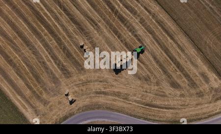 Drohnenbild, das einen Traktor zeigt, der runde Strohballen auf einem kürzlich geernteten Feld nahe dem Gurtenhügel in Bern, Schweiz, transportiert, mit geschwungenen Spuren a Stockfoto