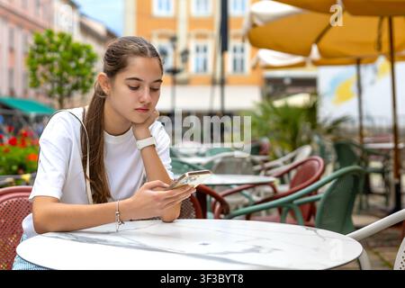 Junge Frau, die ein Smartphone in einem Außencafé benutzt. Junge Frau, die auf ein Smartphone schaut, während sie an einem Marmortisch in einem farbenfrohen Außencafé sitzt. Stockfoto