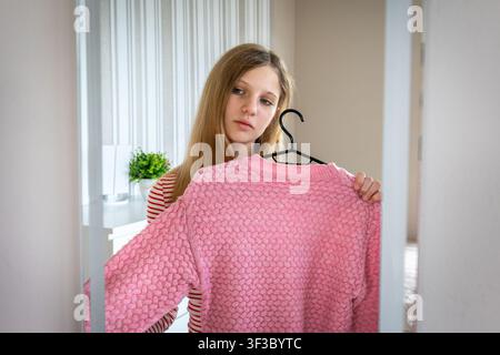 Junge Frau wählt einen rosa Pullover vor einem Spiegel. Junge Frau hält einen rosa gestrickten Pullover auf einem Kleiderbügel, während sie in einen Spiegel im Innenraum schaut. Stockfoto