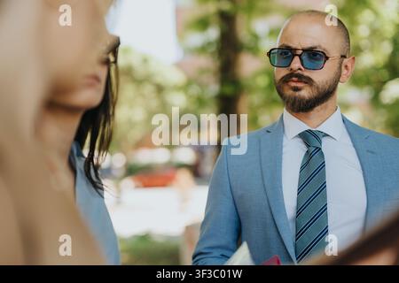 Geschäftsmann in einem blauen Anzug und einer Sonnenbrille, der Kollegen während eines Geschäftstreffens im Freien zuhört Stockfoto
