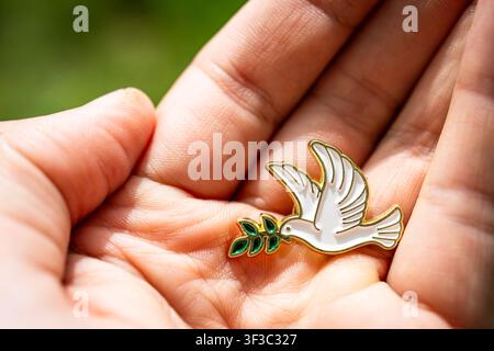 Nahaufnahme einer Emaille-Friedens Taube mit Olivenzweig, die in einer gekrümmten Hand ruht. Bright sun highlights glossy edges while soft green backgr Stockfoto